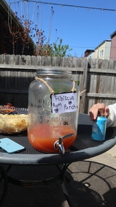 A large glass drink dispenser labelled "Hibiscus Rum Punch" sitting on a a patio table outside