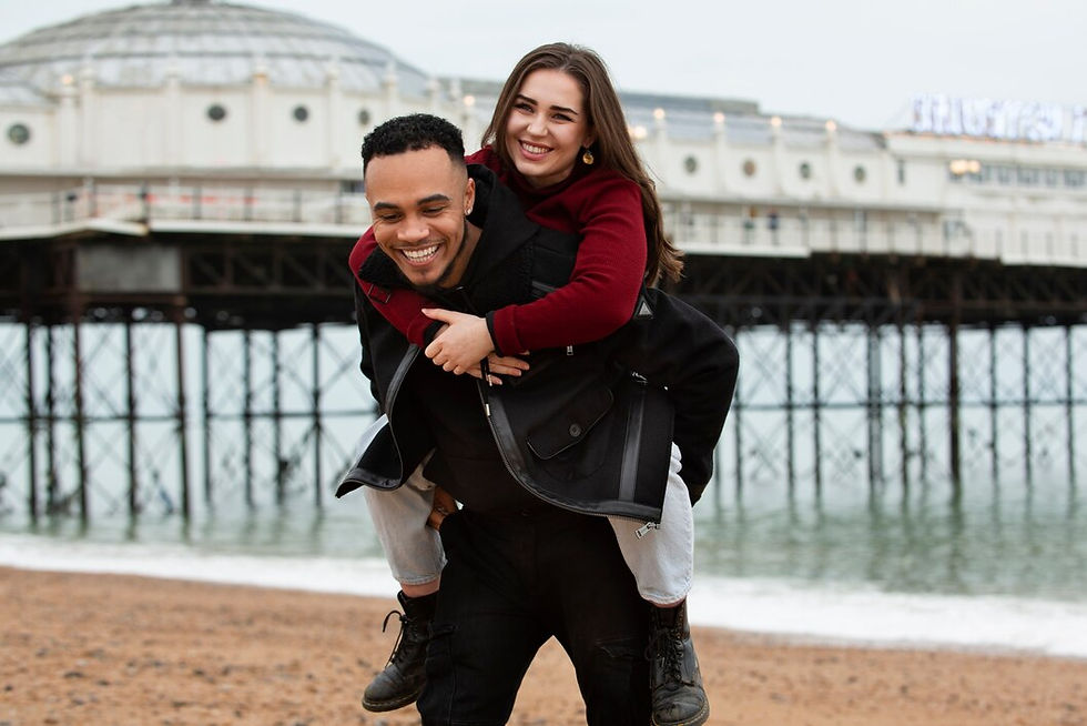 Man piggybacking a woman, both smiling, in front of a pier.