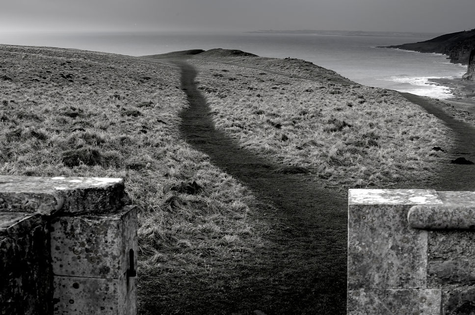 A black and white photograph of a path leading away from the photographer across the top of a cliff covered in thick grass. Beyond the cliff is the sea and sky taking up the top quarter of the image. Part of a low stone gateway can be seen in the image foreground.