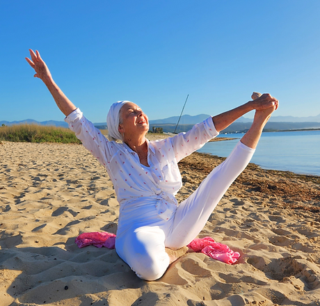 An elder Kundalini Yoga teacher in white on the beach, in a complicated pose, seated with her left leg help up above her head