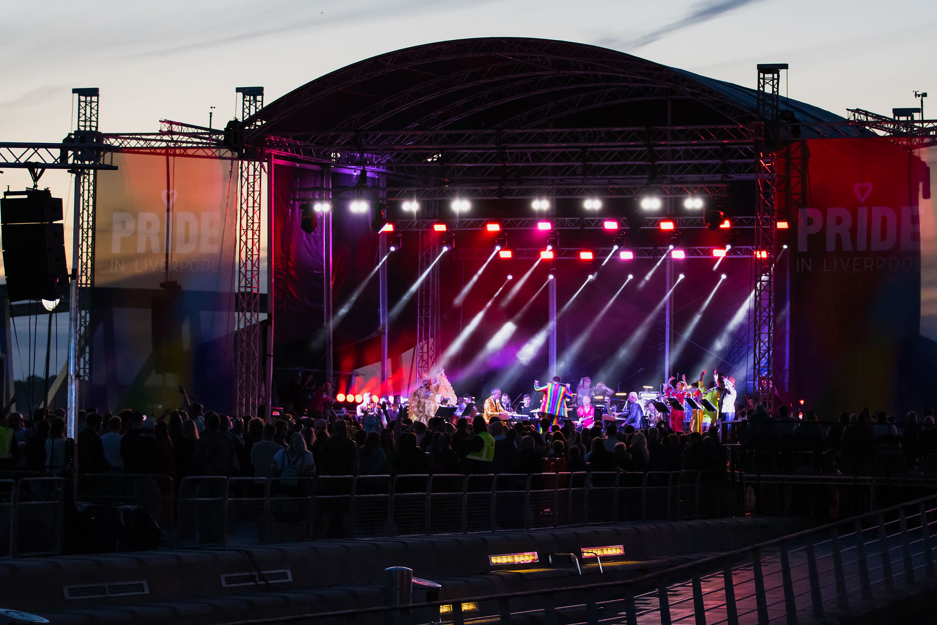 Outdoor stage on the waterfront with orchestra, multi-coloured lighting and audience crowds