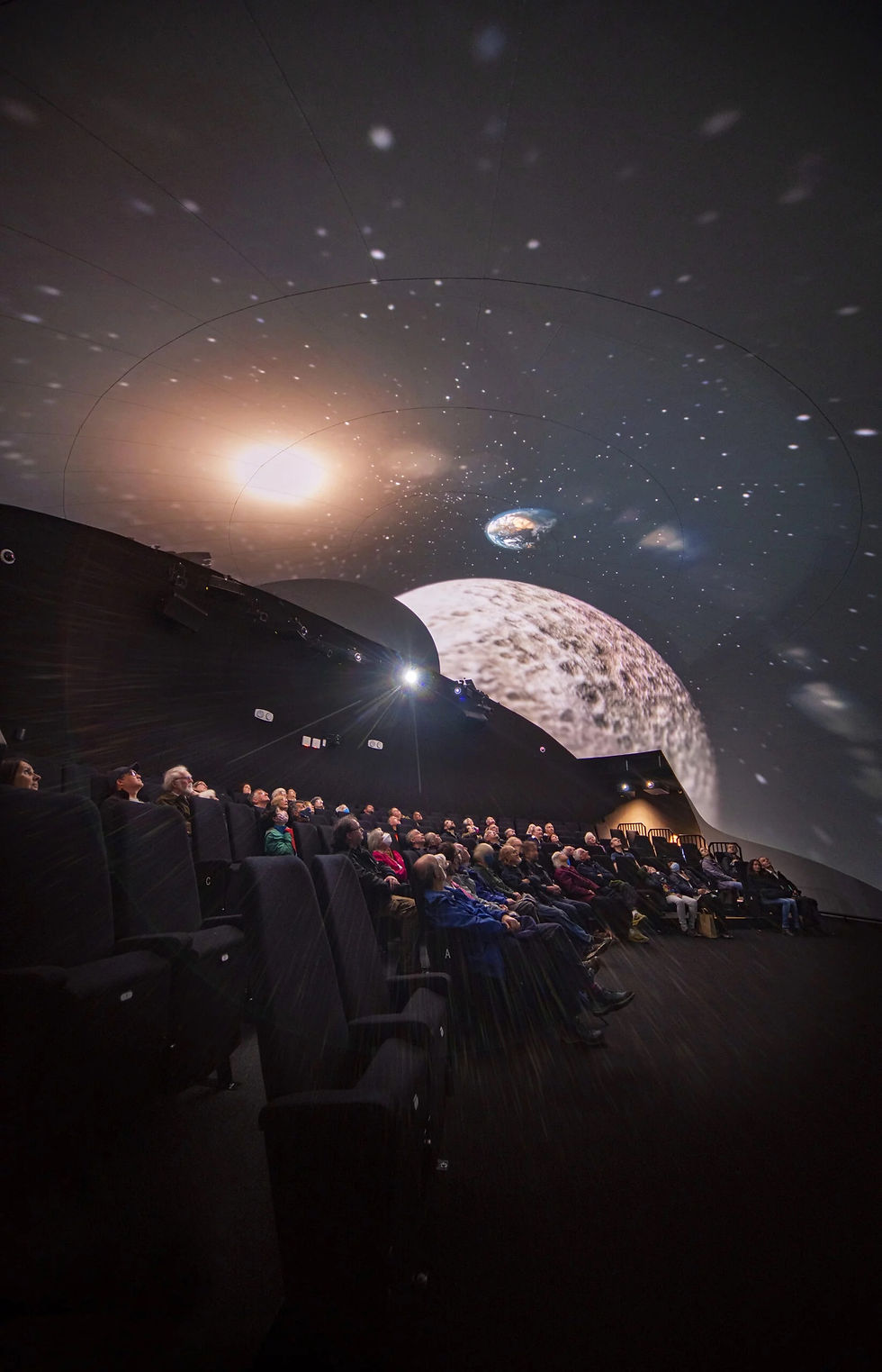 Audience in a dark planetarium watches a projected starry space scene with planets and a bright sun-like glow.