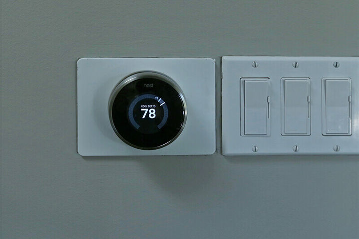 Close-up of a round smart thermostat and a white three-gang light switch plate on a grey wall, Bel Air Manor & Bel Air Cinema.