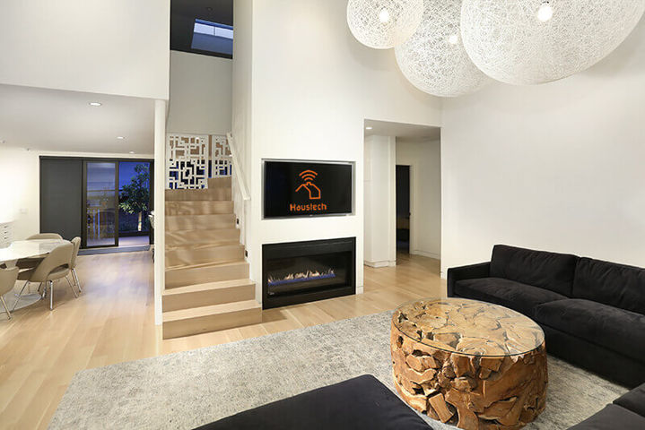 Modern living room featuring a black sofa, rustic wooden coffee table, staircase, and a wall-mounted television above a fireplace, Mar Vista Hilltop Retreat.
