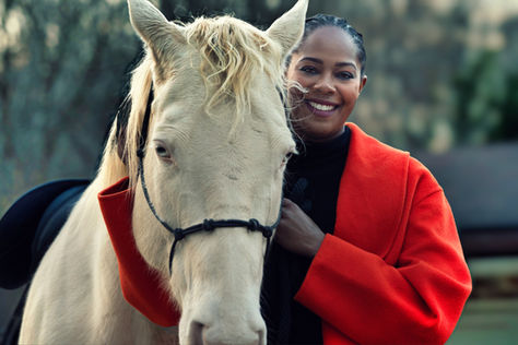 Black woman wearing a red coat standing beside a white horse in an outdoor portrait, holding the reins in a calm, connected moment.