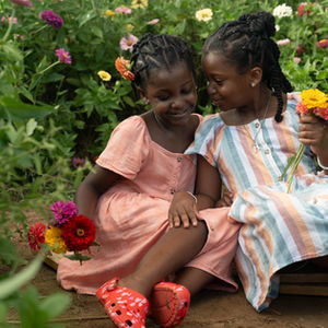 Two young girls sitting together in a colorful flower garden, smiling and holding flowers, captured in a natural light portrait.