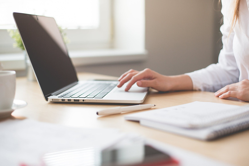 Partial view of a woman sitting infront of a desk touching the touchpad of an open latop. Two notebooks, a pen and a white mug can be seen on the table. There is a blurry view of more notebooks and a cellphone at the front of the image. A white window with natural lighting is observed in the background.