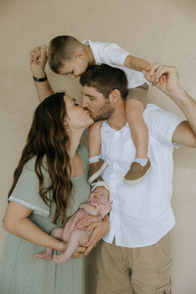 Family of four kissing, with a baby, on shoulders. Families present.