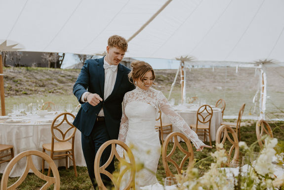 Bride and groom entering wedding reception, smiling, with decorated tables and chairs.