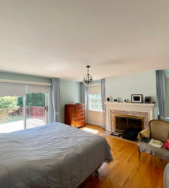 A master bedroom with light blue walls, a large bed with grey bedding, a fireplace, and a sliding glass door leading to a balcony, flanked by custom light blue drapes and Roman shades for privacy and style, by Mill End Shops.