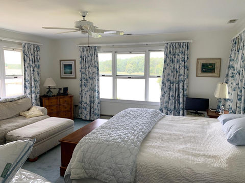 A bright bedroom with multiple windows overlooking a lake, featuring custom blue and white floral patterned drapes, a large bed with white bedding, and a chaise lounge, creating a serene lakeside retreat by Mill End Shops.