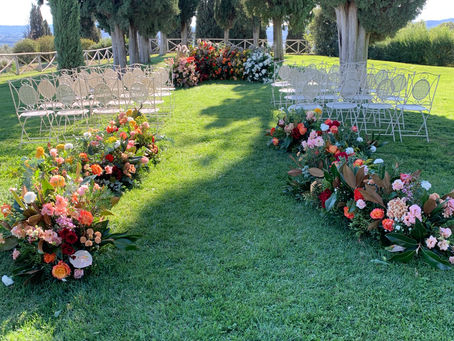 Cerimonia di matrimonio all'aperto a Collemassari in Maremma — composizioni floreali a terra lungo la navata con rose nei toni dell'arancio, pesca e bordeaux, cipressi e paesaggio toscano sullo sfondo