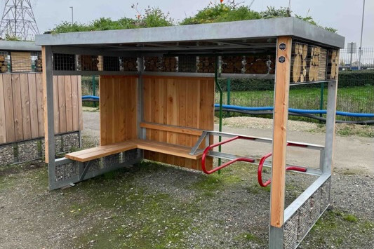A green roof shelter with a bnch and cycle parking built in. Plants grow on the roof and there is some timber cladding and corner posts softening the look of the steel frame.