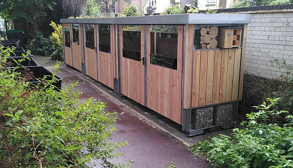 Modular euro bin store units layed out in a row. These bin shelters have a rich slections of plants growing on the roof, habitat panels and bird boxes on the sides. The stores are clad in timber with stainless steel bin access slots in the doors.