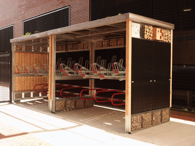 Two shelters side by side offer cycle parking using two tier or double height cycle racks. The structure is part of a green roof shelter with a growing roof and wildlife habitat on its facade. One end is UK Larch clad, the other uses twin-mesh.
