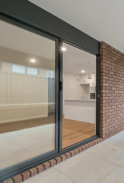 Photo taken from an undercover alfresco showing a tiled floor, brown brick house, looking down the side of the house to a grassed area in the backyard and on the left into the kitchen and dining room of the house through a large glass sliding door.