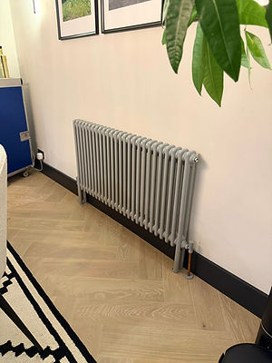 Gray column radiator installed on white wall with light wood flooring.