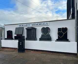 Outdoor display of memorial headstones against a white wall under a "Moriarty Memorials" sign