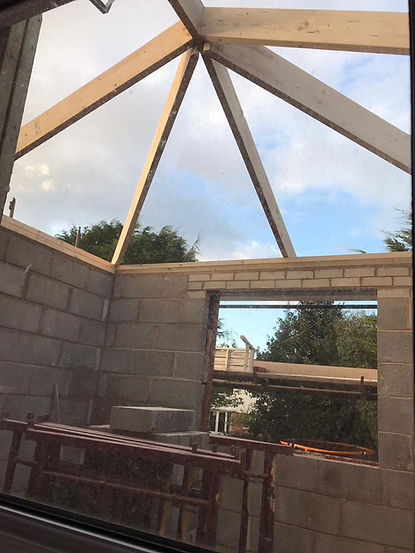 View of a building under construction, featuring a partly built brick wall and exposed triangular roof beams