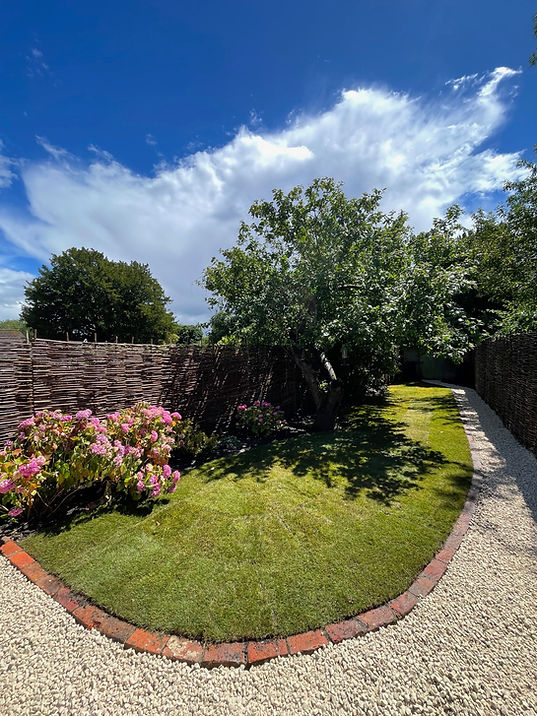 Curved brick-edged lawn with pink flowers, gravel path, and wicker fence under blue sky