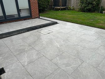 Grey porcelain patio tiles, lawn, brick wall, and a window frame.