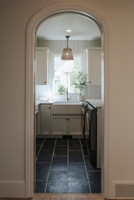Elegant laundry room with white cabinets, farmhouse sink, dark stone tile, and an arched doorway.