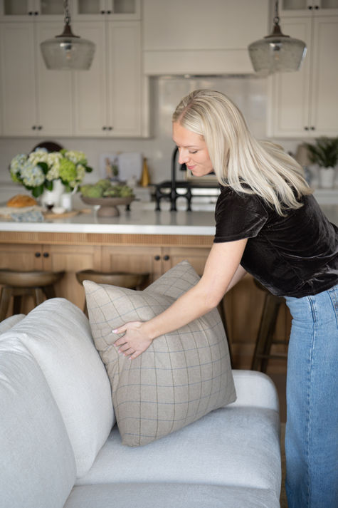 A woman in a black t-shirt fluffing a grey pillow on a sofa by the kitchen.