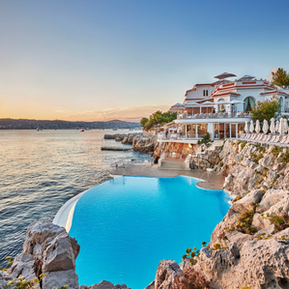 Aerial view of Hôtel du Cap Eden Roc in Antibes at sunset, white cliffside property with an infinity pool carved into the rocks above the Mediterranean.