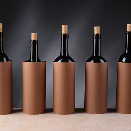 Five wine bottles wrapped in plain brown paper sleeves concealing their labels, arranged in a row against a dark grey background.