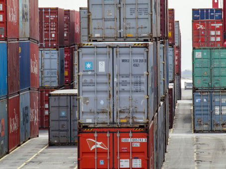 Stacks of Black-owned colorful shipping containers in a port, with visible labels and numbers, creating a geometric pattern against a cloudy sky backdrop.