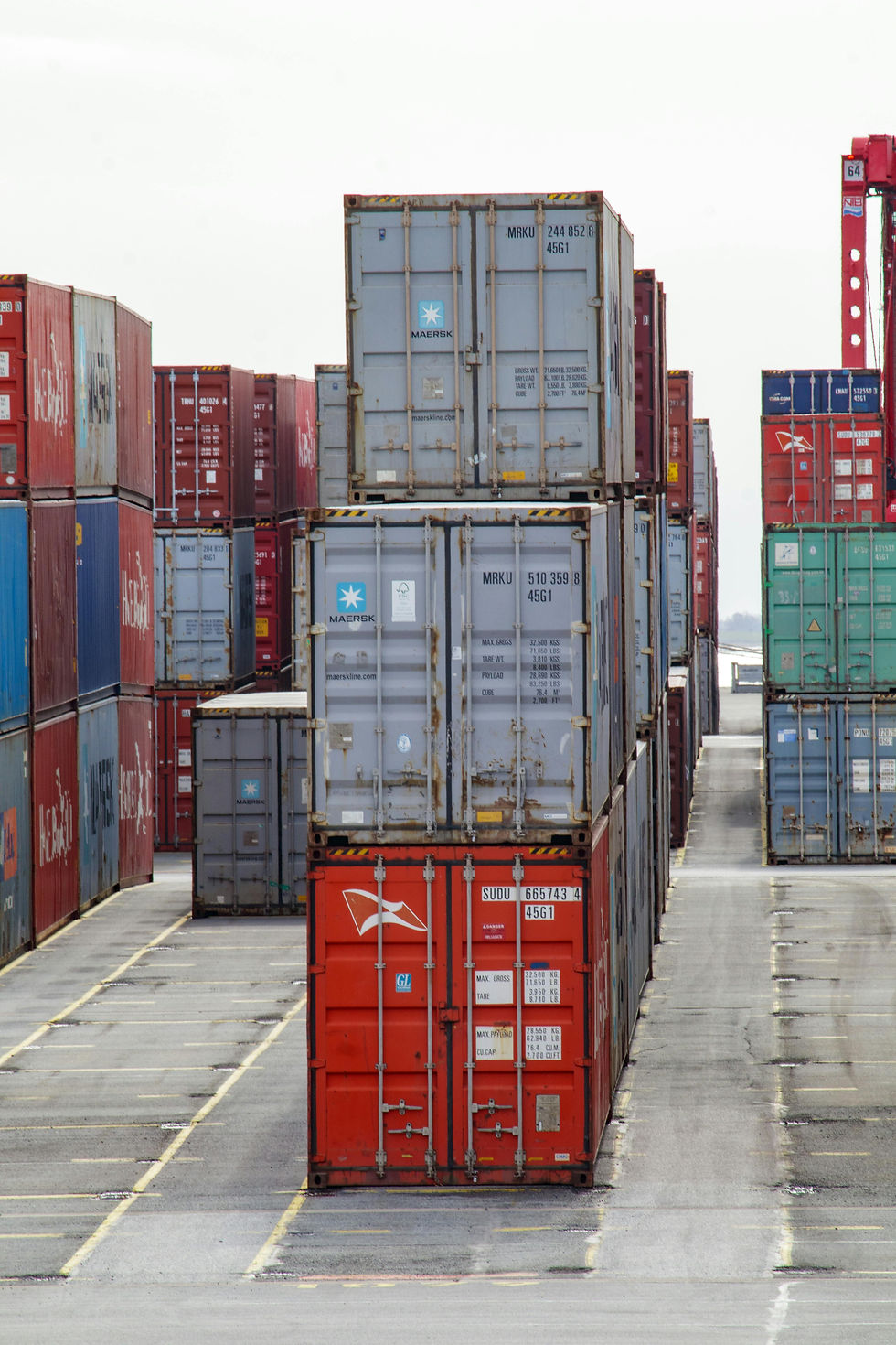 Stacks of Black-owned colorful shipping containers in a port, with visible labels and numbers, creating a geometric pattern against a cloudy sky backdrop.