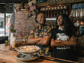 Smiling Black man and Black woman with crossed arms at a bar, surrounded by bottles and drinks, with brick wall background and a casual vibe.