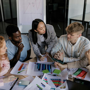 Five people in business attire discuss paperwork with graphs in a meeting room. A chart is visible on a whiteboard. Engaged, focused mood.