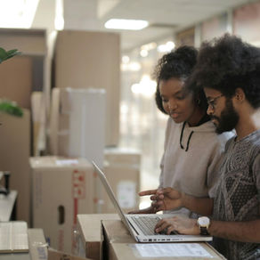 Two people using a laptop in a room with cardboard boxes and plants. They appear focused on the computer screen . Bright lighting and blurred background.