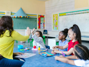 Teacher high-fives a child in a colorful classroom, while black child watches. Children sit at a blue table with toys, smiling and engaged. Chalkboard in background.