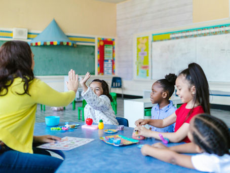 Teacher high-fives a child in a colorful classroom, while black child watches. Children sit at a blue table with toys, smiling and engaged. Chalkboard in background.