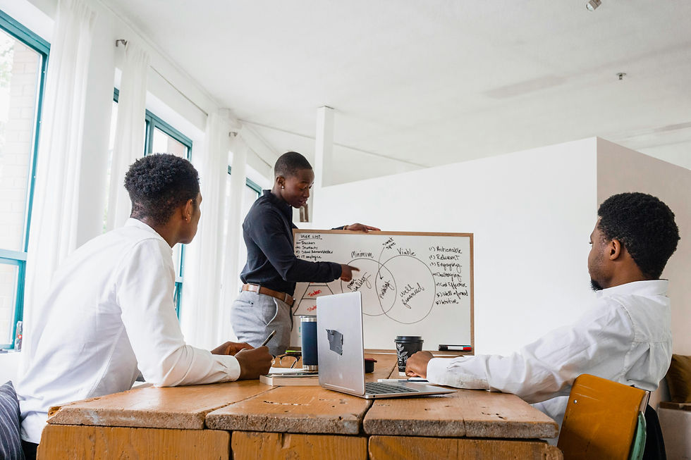 Three Black men in a meeting room, one presenting on a whiteboard with diagrams. Others seated at a wooden table, engaged. Bright atmosphere.