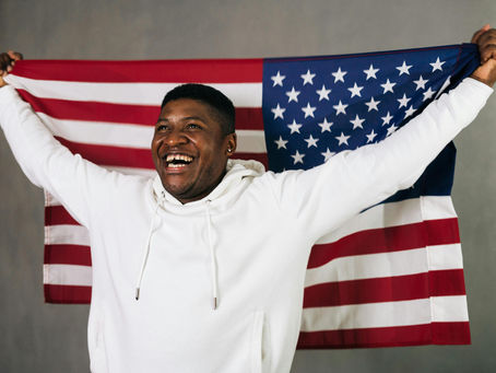 Man in white hoodie joyfully holds up a large American flag. Gray background highlights the red, white, and blue colors of the flag.