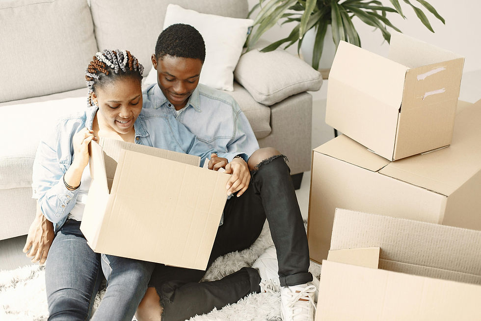 A couple unpacks moving boxes in a bright living room, sitting on a rug near a couch. Green plant in the background adds freshness.