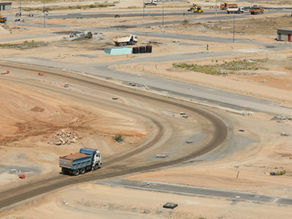 Dump truck on dusty African construction site, driving on curved dirt road. Sparse buildings and equipment in background, under sunny sky.