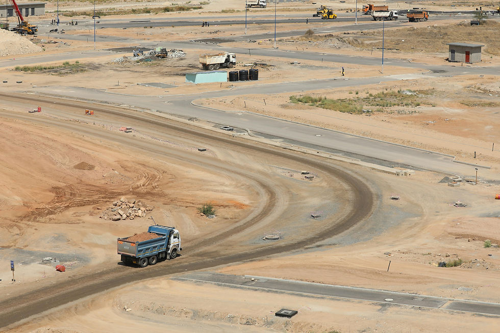Dump truck on dusty African construction site, driving on curved dirt road. Sparse buildings and equipment in background, under sunny sky.