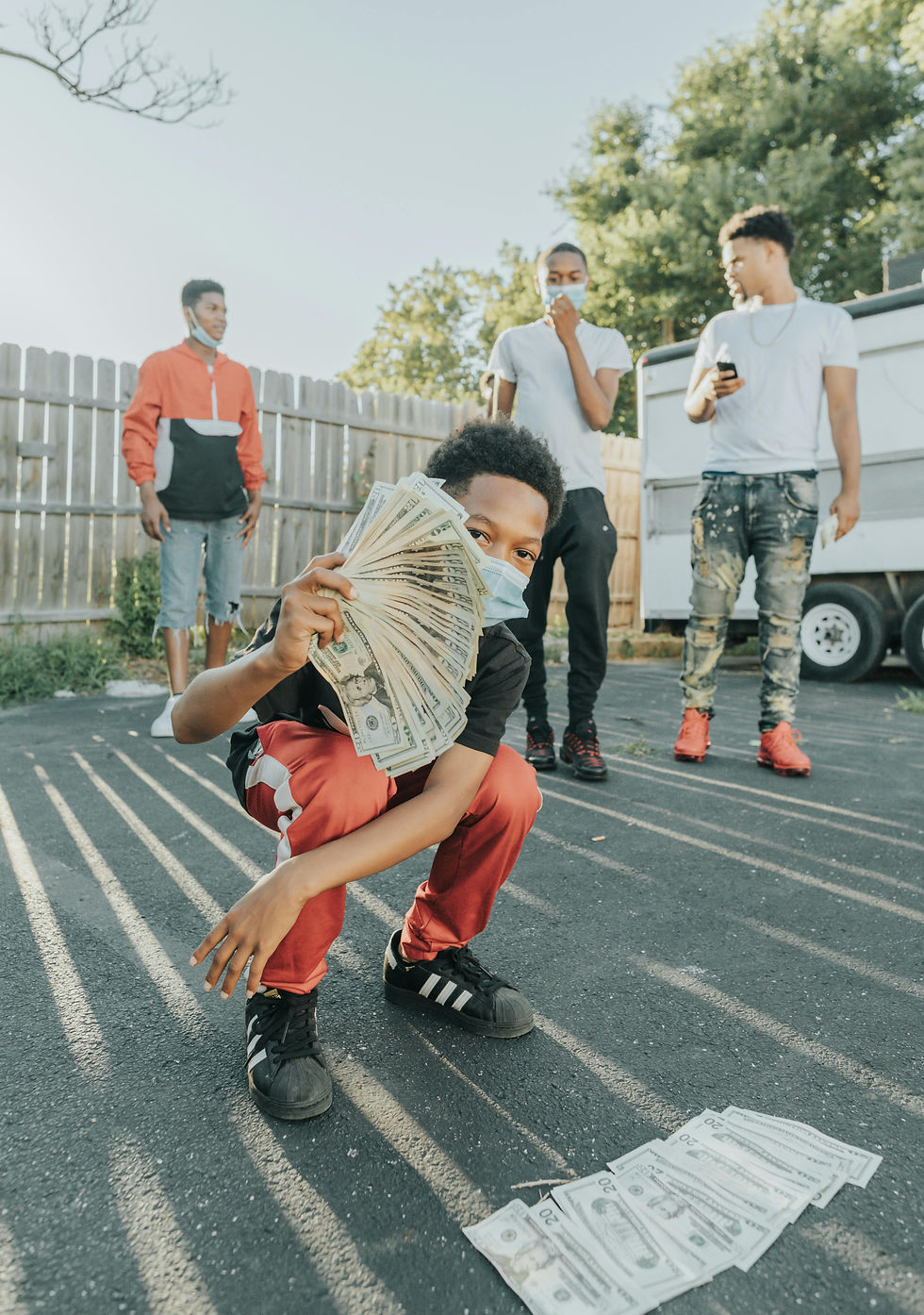A child in a mask crouches, holding cash and waving it in front of the camera, with three teens in casual wear standing behind on a sunlit paved area, near a wooden fence.