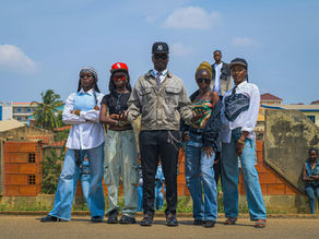 Five people in urban attire stand confidently on a street. Background: palm trees, buildings, blue sky. Casual and stylish vibe.