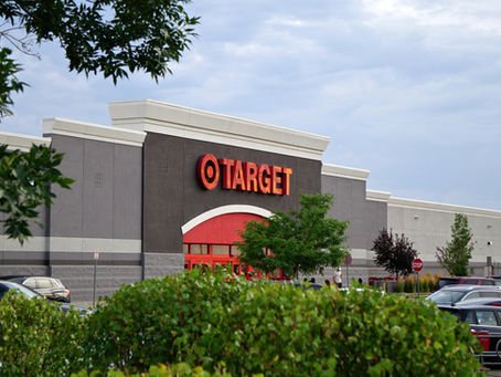 Target store exterior with red logo and entrance. Cars parked in the lot, greenery in foreground, cloudy sky above.