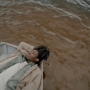 Man in beige coat relaxes on boat, hands behind head, with closed eyes. Calm water in the background; a sense of tranquility pervades.