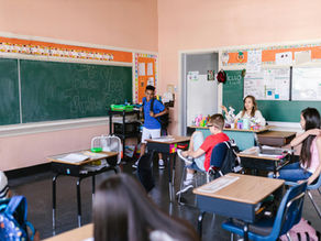 Classroom scene: a Black student stands smiling with a backpack, teacher seated at desk, students around, with soft focus on Black student