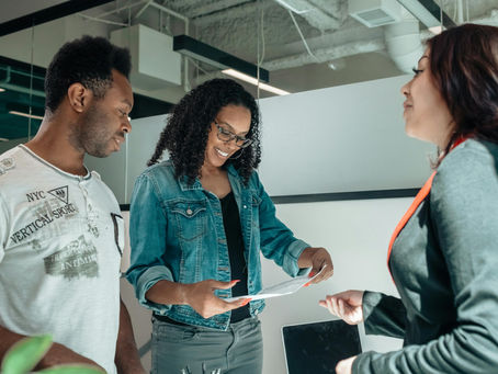 Three people in an office, one holding a document. They're smiling and engaged in conversation. Married couple smiling and looking at a document.