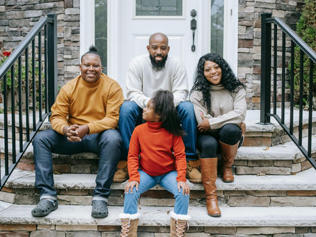 Black family of four sitting on stone steps, smiling. Casual attire in earthy tones. Brick facade and railing in background. Relaxed, happy mood.