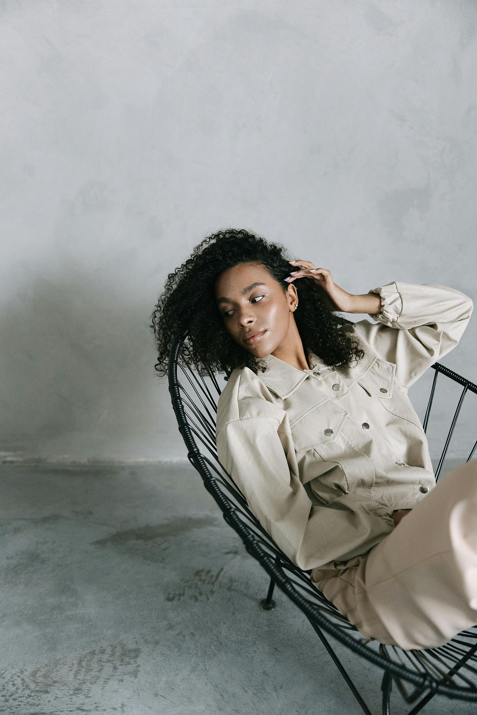 Person reclines on a black wire chair, wearing a beige jacket, relaxed expression. Gray concrete wall background, minimalist setting.