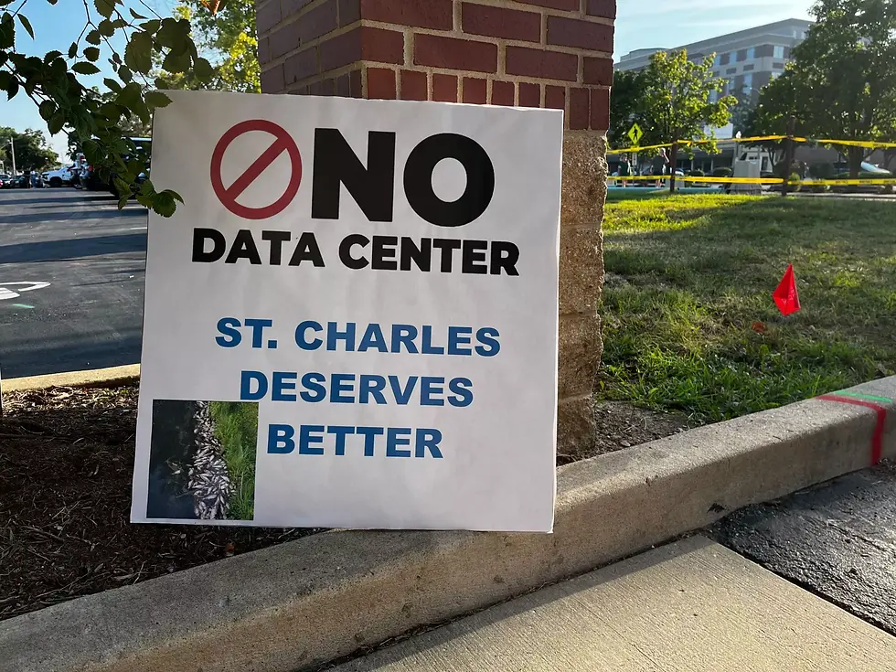 Sign reading "No Data Center, St. Charles Deserves Better" placed near a brick wall with trees in the background, conveying protest mood.